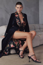 Load image into Gallery viewer, A fashion shot of a woman wearing a black velvet full length robe with contrasting floral silk lapel, cuffs and lining. It is tied at the waist with a belt – one side velvet, the other silk. A contrast blush pink piping finishes the trims, and sleeves.