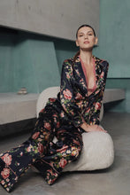 Load image into Gallery viewer, A woman wearing Benet pyjamas made of stretch silk. The fabric is a fabulous floral print on a black background. The flowers are a soft dusky pink colour and gentle green leaves finish the motif.
Contrast piping (also in dusky pink) is located about 3 inches from the bottom of the pant leg, on the edging of the top and lapel and bottom of the sleeves.