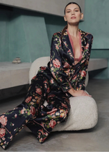Load image into Gallery viewer, A woman wearing Benet pyjamas made of stretch silk. The fabric is a fabulous floral print on a black background. The flowers are a soft dusky pink colour and gentle green leaves finish the motif.
Contrast piping (also in dusky pink) is located about 3 inches from the bottom of the pant leg, on the edging of the top and lapel and bottom of the sleeves.