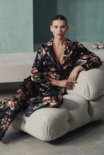 Load image into Gallery viewer, A woman wearing Benet pyjamas made of stretch silk. The fabric is a fabulous floral print on a black background. The flowers are a soft dusky pink colour and gentle green leaves finish the motif.
Contrast piping (also in dusky pink) is located about 3 inches from the bottom of the pant leg, on the edging of the top and lapel and bottom of the sleeves.