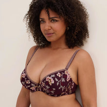 Load image into Gallery viewer, A woman with curly dark hair wearing a dark burgundy floral embroiders bra with embroidered straps, photographed against a plain off-white wall.