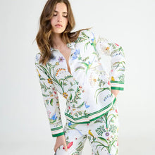 Load image into Gallery viewer, A woman modeling a matching two-piece pyjama set with a white base and a colorful botanical print featuring green leaves, blue flowers, and green striped accents on the cuffs and hem.