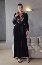 Load image into Gallery viewer, A woman wearing a black velvet full lenth robe with contrasting floral silk lapel, cuffs and lining. It is tied at the waist with a belt – one side velvet, the other silk. A contrast blush pink piping finishes the trims, and sleeves.
