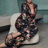A woman wearing Benet pyjamas made of stretch silk. The fabric is a fabulous floral print on a black background. The flowers are a soft dusky pink colour and gentle green leaves finish the motif.
Contrast piping (also in dusky pink) is located about 3 inches from the bottom of the pant leg, on the edging of the top and lapel and bottom of the sleeves. 
