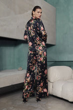 Load image into Gallery viewer, A woman wearing Benet pyjamas made of stretch silk. The fabric is a fabulous floral print on a black background. The flowers are a soft dusky pink colour and gentle green leaves finish the motif.
Contrast piping (also in dusky pink) is located about 3 inches from the bottom of the pant leg, on the edging of the top and lapel and bottom of the sleeves. 


