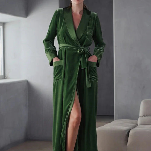 A woman wearing an Ivy green silk velvet full length dressing gown. It is tied at the waist with a belt that has a satin side. The pockets are at the front with a satin trim including a statement breast pocket. The lapel and cuffs also are finished in satin silk.