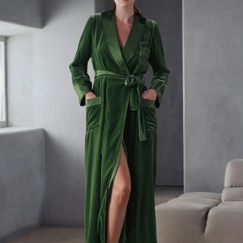A woman wearing an Ivy green silk velvet full length dressing gown. It is tied at the waist with a belt that has a satin side. The pockets are at the front with a satin trim including a statement breast pocket. The lapel and cuffs also are finished in satin silk.