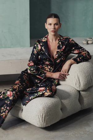 A woman wearing Benet pyjamas made of stretch silk. The fabric is a fabulous floral print on a black background. The flowers are a soft dusky pink colour and gentle green leaves finish the motif.
Contrast piping (also in dusky pink) is located about 3 inches from the bottom of the pant leg, on the edging of the top and lapel and bottom of the sleeves. 
