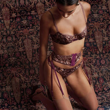 Load image into Gallery viewer, A woman kneels on a patterned oriental rug wearing a matching lingerie set with a metallic burgundy and gold pattern, sheer thigh-high stockings, and a purple lace detail on the waistband.
