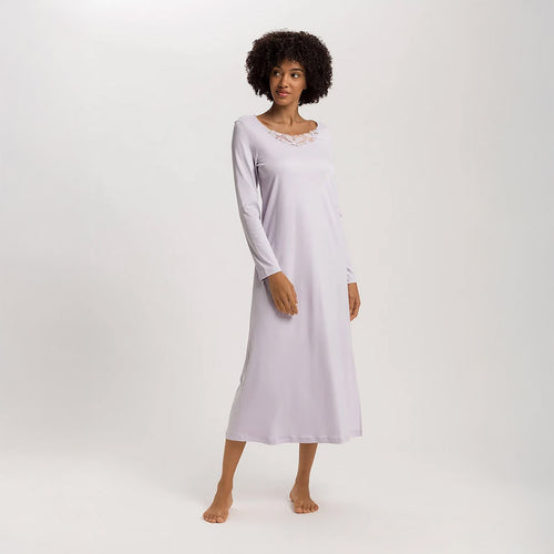 A woman wearing a Pink Lavender long sleeve full length (130cm) nightdress. It has a lovely round neckline embellished with a delicate embroidered lace trim on tulle. The garment has a lovely feminine flow to it with a slit on the side to allow for comfortable movement.