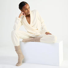 Load image into Gallery viewer, A model sits on a white block wearing a cream-colored lounge set consisting of a shawl-collar bed jacket paired with beige ribbed socks against a plain white studio background.
