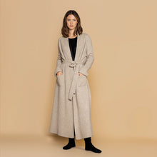 Load image into Gallery viewer, A model wearing a full length cashmere dressing gown in biscuit colour.  her hands are in Square  pockets at the front. Belt tied around the waist.
