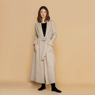 A model wearing a full length cashmere dressing gown in biscuit colour.  her hands are in Square  pockets at the front. Belt tied around the waist.