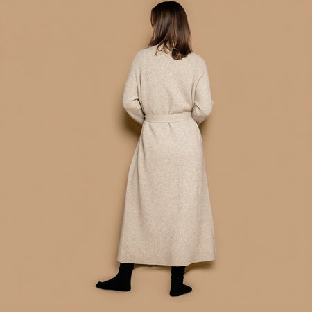 A woman viewed from behind wearing a long, light beige cashmere knitted coat with a matching belt, paired with black socks, against a solid beige background.