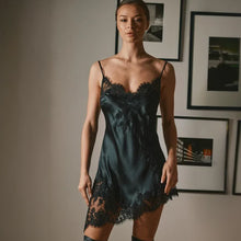 Load image into Gallery viewer, A woman wearing a black silk satin slip dress with asymmetrical black lace trim, standing in a room with framed wall art.
