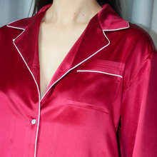 Load image into Gallery viewer, A close-up of a person wearing a satin pajama top in deep red with thin white piping along the collar, placket, and chest pocket.
