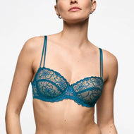 A woman wearing a teal blue lace underwire bra against a plain light gray background.