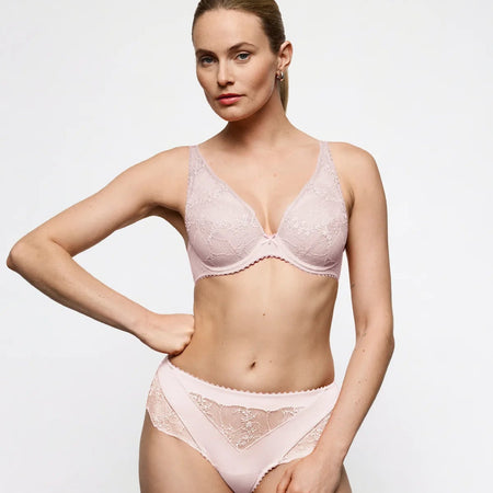A woman modeling a light pink lace bra and matching high-waisted briefs against a plain light gray studio background.