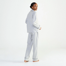 Load image into Gallery viewer, A model wearing a light gray two-piece pajama set with dark piping, standing barefoot against a clean white studio background.
