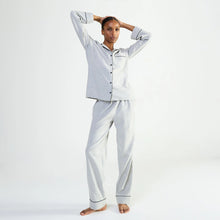 Load image into Gallery viewer, A woman modeling a light gray two-piece pajama set with dark piping details, consisting of a long-sleeve button-up shirt and matching pants, against a plain white studio background.
