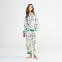 Load image into Gallery viewer, A woman modeling a matching two-piece pyjama set with a white base and a colorful botanical print featuring green leaves, blue flowers, and green striped accents on the cuffs and hem.
