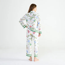 Load image into Gallery viewer, A woman stands with her back to the camera wearing a matching long-sleeve pajama set with a white base and a colorful botanical and bird pattern, featuring green striped cuffs and hem.
