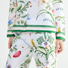Load image into Gallery viewer, A close-up of a model wearing a matching two-piece pajama set with a white base and a colorful botanical and bird print. The set includes a long-sleeve top and pants, featuring green and pink striped accents along the hem and cuffs, with yellow birds and green leaves throughout.
